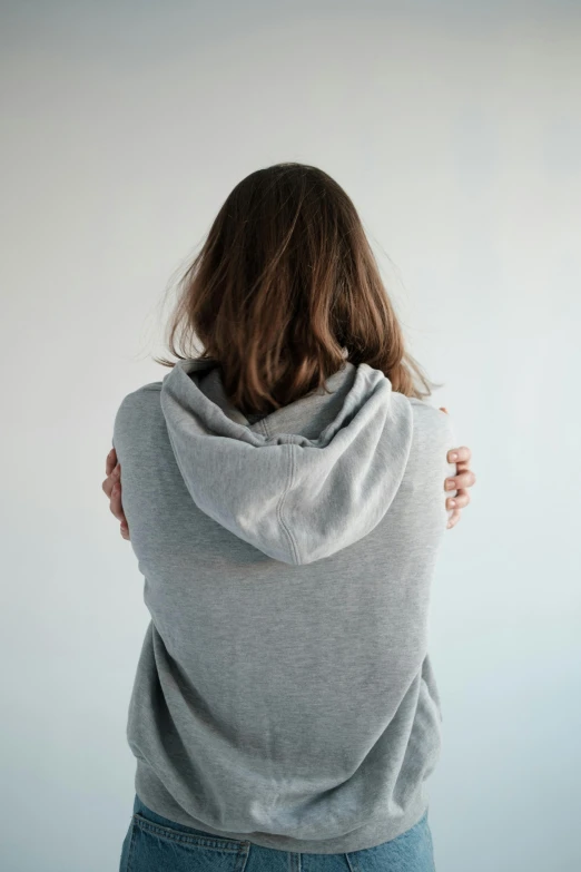 a woman standing with her back to the camera, happening, light grey hoodie opened, arm around her neck, light grey backdrop, wearing ragged clothing