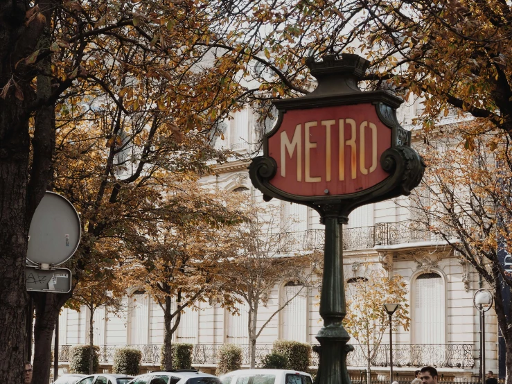 a red metro sign sitting on the side of a road, by Meredith Dillman, trending on unsplash, art nouveau, splendid haussmann architecture, autumn season, square, fan favorite