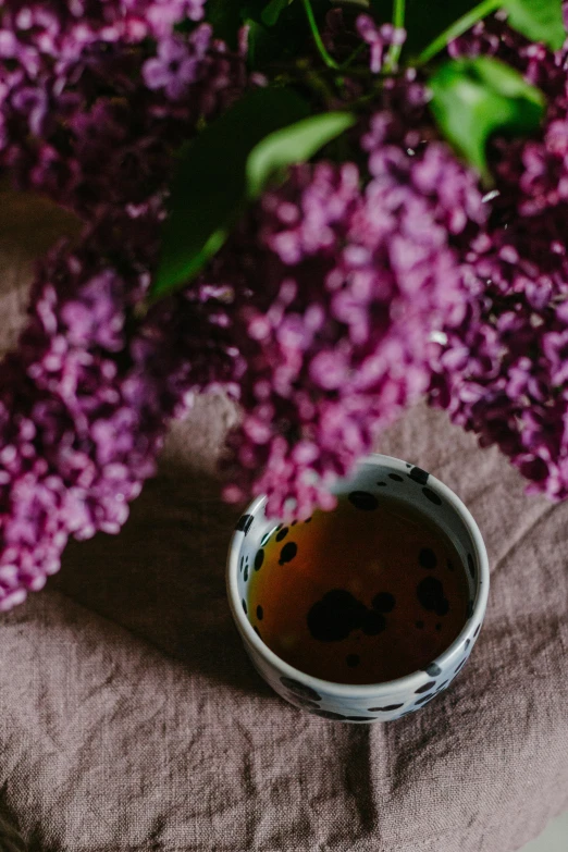 a cup of tea next to a bunch of purple flowers, a still life, unsplash, close up of lain iwakura, linen, patterned, wine