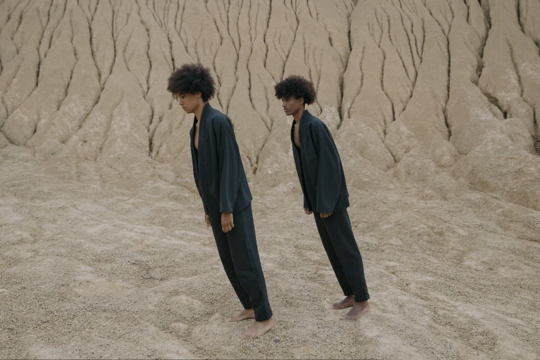 a couple of people that are standing in the sand, an album cover, by Caro Niederer, unsplash, conceptual art, jumpsuit, twins, wearing dark green robes, a portrait of issey miyake