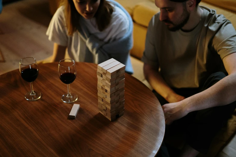 a man and a woman sitting at a table with wine glasses, jenga tower, blockout, playing games, wooden coffee table