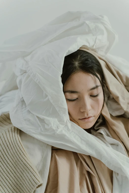 a woman laying on top of a bed under a blanket, inspired by Elsa Bleda, unsplash contest winner, visual art, asian woman made from origami, model wears a puffer jacket, veiled face, white cloak