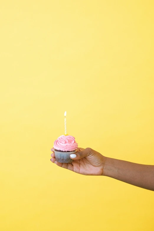 a person holding a cupcake with a candle in it, by Winona Nelson, trending on unsplash, photorealism, pink and yellow, happy birthday, dwell, essence