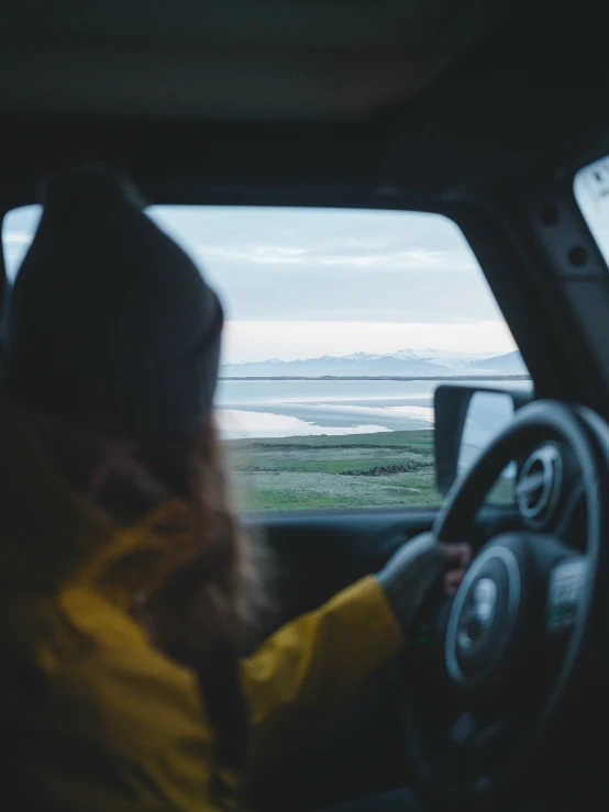 a woman sitting in the driver's seat of a car, by Christen Dalsgaard, unsplash contest winner, looking out at the ocean, wearing a yellow hoodie, eerie moorlands behind her, 🚿🗝📝