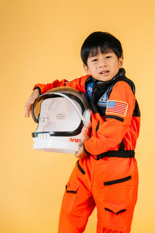 a young boy in an orange space suit holding a helmet, pexels contest winner, light and space, asian descent, pilot outfit, full product shot, science magazine cover