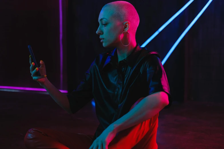 a man sitting on the ground holding a cell phone, an album cover, inspired by Elsa Bleda, pexels contest winner, antipodeans, asian nymph bald goddess, nightclub dancing inspired, shaved hair, red neon