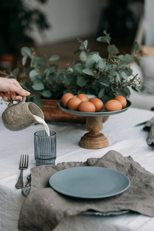 a person pouring water into a cup on a table, a still life, inspired by Constantin Hansen, trending on unsplash, eggs, eucalyptus, festive atmosphere, grey and blue theme