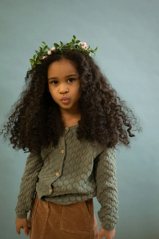 a little girl with a flower crown on her head, inspired by Kate Greenaway, renaissance, long wild black curly hair, at a fashion shoot, wearing a green sweater, textured