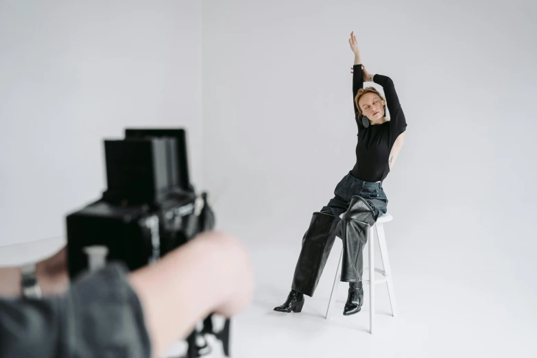 a woman sitting on a stool in front of a camera, trending on pexels, arms stretched out, wearing in black clothes, person in foreground, shooting pose