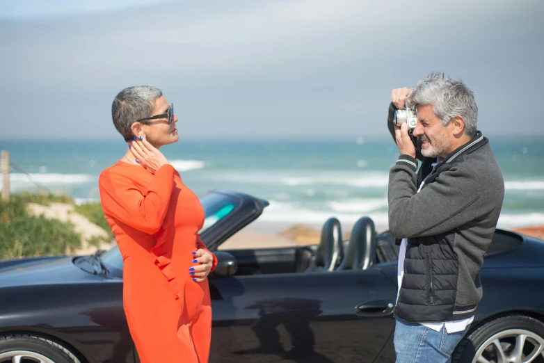 a man standing next to a woman in front of a car, by David Donaldson, pexels contest winner, philippe starck, standing near the beach, behind the scenes, jeff goldblum