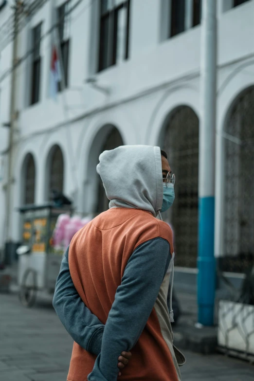 a person wearing a face mask walking down a street, trending on pexels, renaissance, gray hoodie, back of head, nepal, grey orange