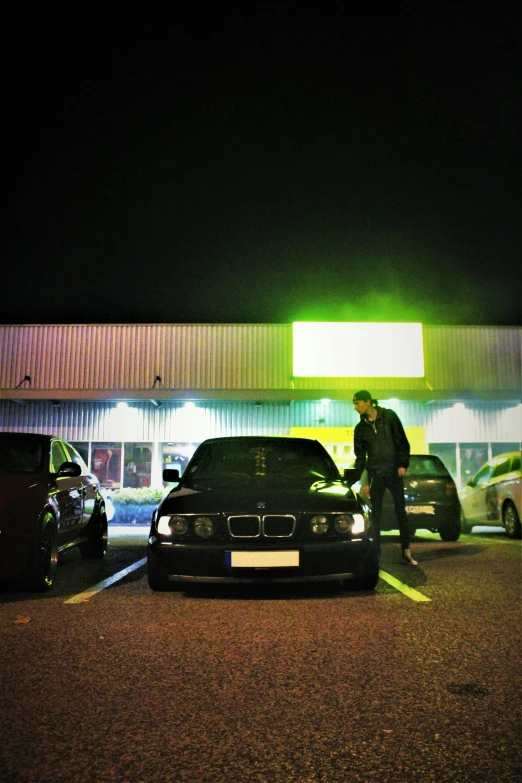 a man standing next to a car in a parking lot, an album cover, by Tom Bonson, renaissance, night clubs and neons, bmw e 3 0, taken in the late 2010s, chillin at the club together