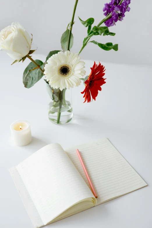 a vase of flowers and a notebook on a table, lit candles, set against a white background, contemplating, lined paper