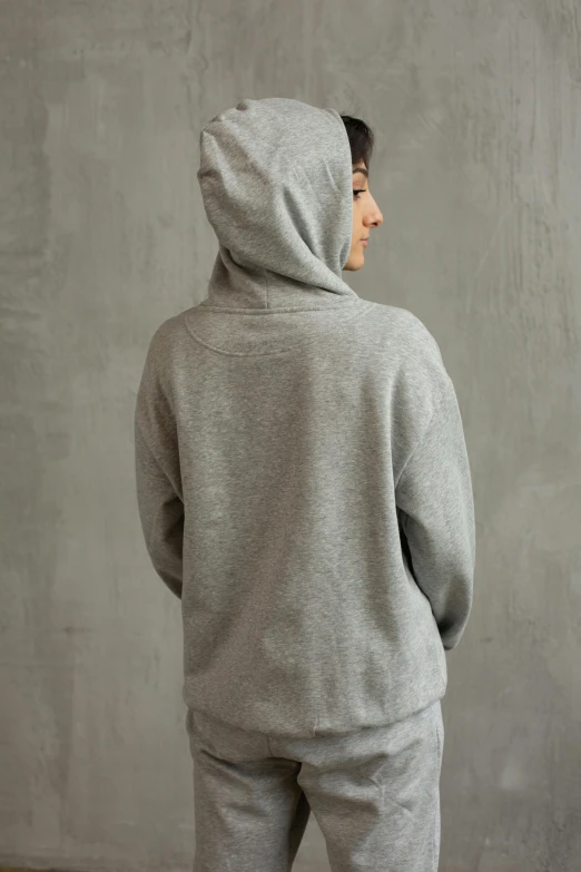 a man standing in front of a wall wearing a hoodie, inspired by Judy Takács, showing her shoulder from back, wearing a grey hooded sweatshirt, environmental shot, medium - shot