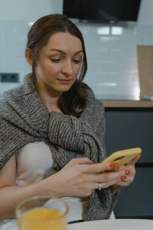 a woman sitting at a table using a cell phone, covered with blanket, wearing a sweater, grey, cooked