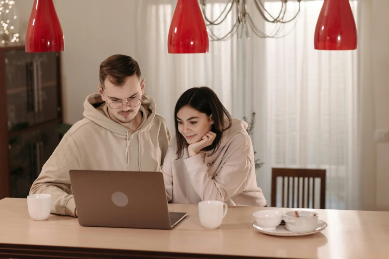 a man and a woman sitting at a table with a laptop, a picture, trending on pexels, wearing a scarlet hoodie, brown, sitting across the room, kacper niepokolczycki