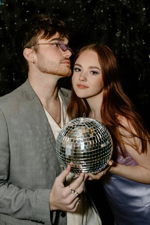 a man standing next to a woman holding a disco ball, an album cover, by Julia Pishtar, trending on pexels, ginger hair with freckles, sophie turner, couple pose, at the party