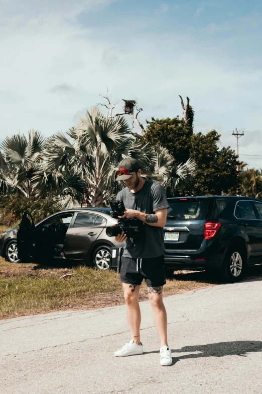 a man standing on the side of a road with a skateboard, kicking a florida mansion, holding a big camera, chicken on top of a car, drone photo