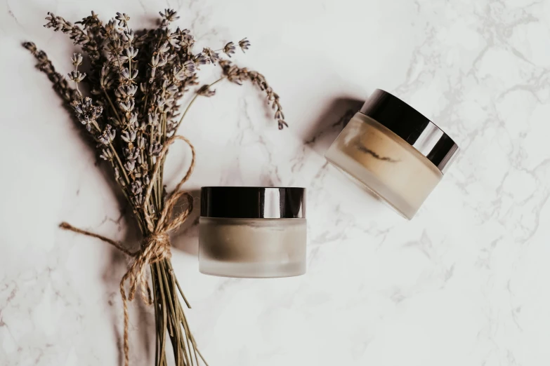 two jars of cream next to a bunch of dried flowers, trending on pexels, pronounced facial contouring, lavender plants, silver，ivory, background image