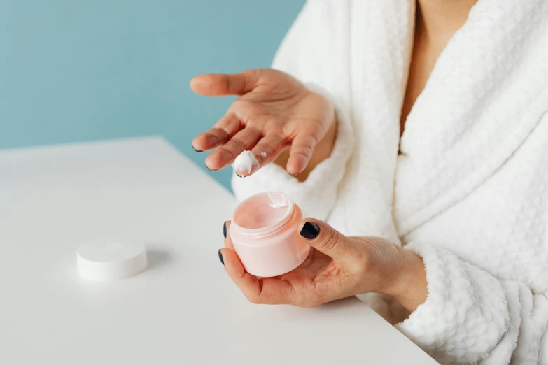 a woman in a bathrobe holding a jar of cream, a picture, by Emma Andijewska, trending on pexels, pink white turquoise, partially cupping her hands, on a white table, skincare