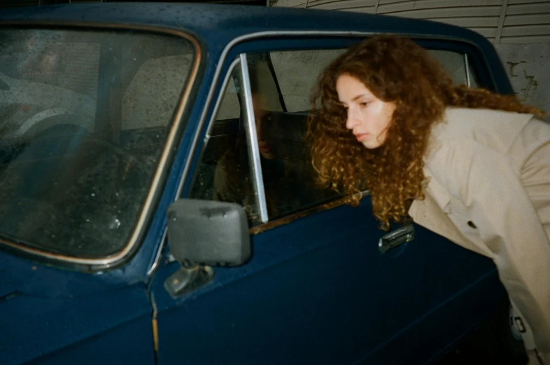 a woman leaning over the window of a blue car, inspired by Nan Goldin, curly brown hair, petra collins and mc. escher, moskvich, garage