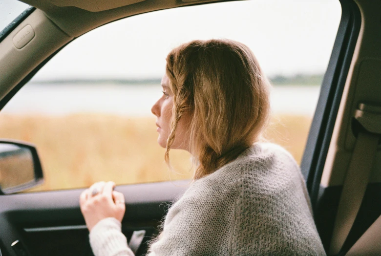 a woman sitting in the driver's seat of a car, trending on unsplash, visual art, sydney sweeney, wearing a cardigan, gazing at the water, side profile view