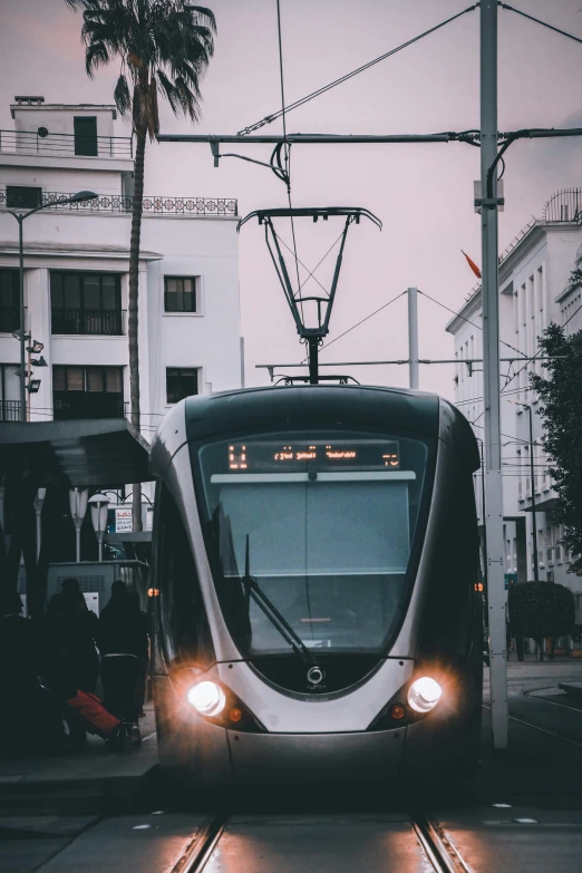 a train traveling down a city street next to tall buildings, by Matija Jama, unsplash contest winner, futuristic marrakech morocco, dark city bus stop, magnificent oval face, black wired cables