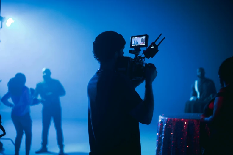 a man holding a camera in front of a group of people, pexels, video art, cinematic blue lighting, movie set”, ( ( theatrical ) ), behind the scenes