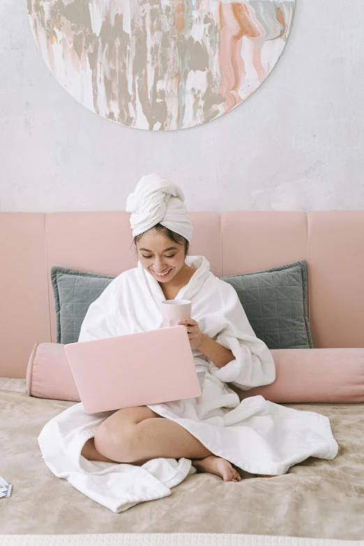 a woman sitting on a bed using a laptop, pexels contest winner, happening, pastel pink robes, skincare, girl with messy bun hairstyle, wearing a towel