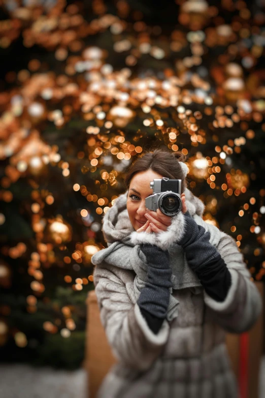 a woman taking a picture in front of a christmas tree, by Adam Marczyński, grey, golden glow, holding a big camera, bokeh ”