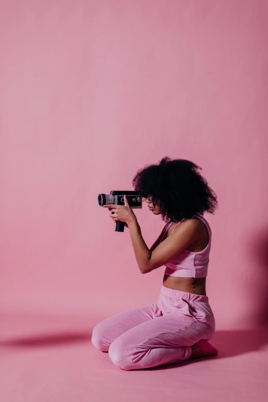 a woman sitting on the ground holding a camera, by Dulah Marie Evans, afrofuturism, brightly lit pink room, shooting a gun, focus her back, solid background