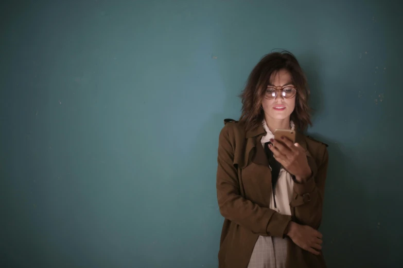 a woman standing in front of a blue wall holding a cell phone, an album cover, pexels, happening, with square glasses, wearing green jacket, brown haired, standing in a dimly lit room