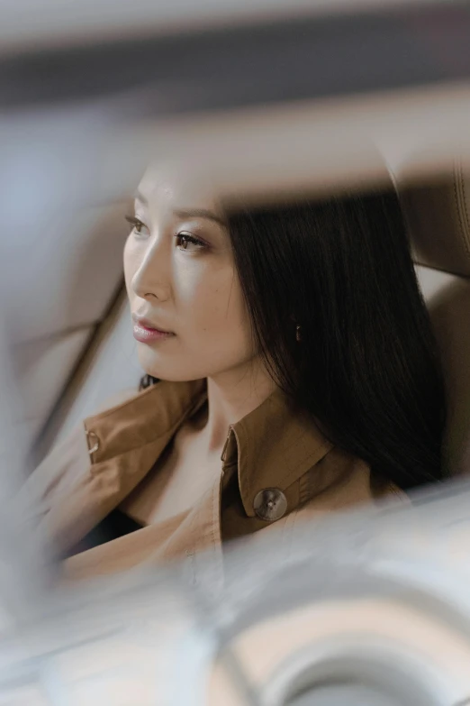 a woman sitting in a car looking out the window, inspired by Li Di, lucy liu portrait, wearing a long beige trench coat, bo chen, contemplative