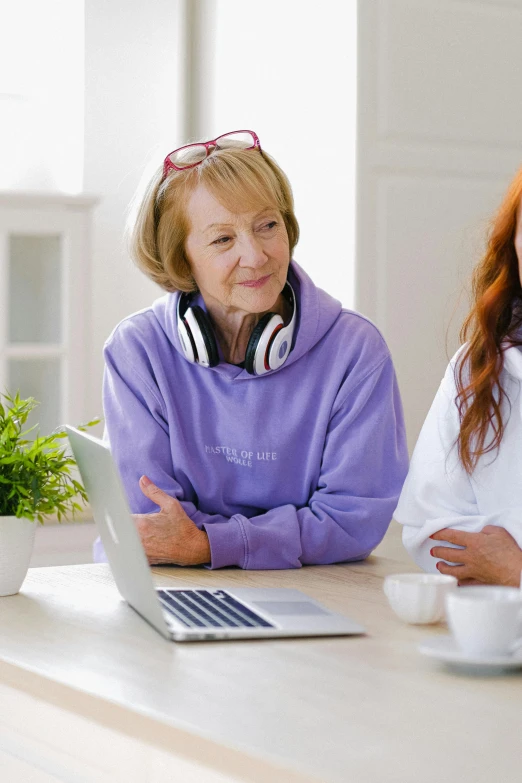 two women sitting at a table with a laptop, wearing a purple sweatsuit, with headphones, wearing a robe, mary jane ansell