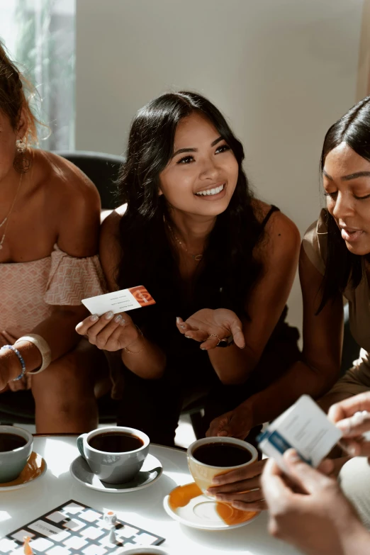 a group of women sitting around a table playing a board game, trending on pexels, happening, photoshoot for skincare brand, sitting on a mocha-colored table, holding an ace card, one single tribe member