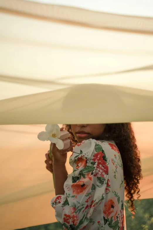 a woman holding a flower up to her face, by Alexis Grimou, trending on unsplash, renaissance, desert robe, under large green umbrella, tanned ameera al taweel, at a fashion shoot