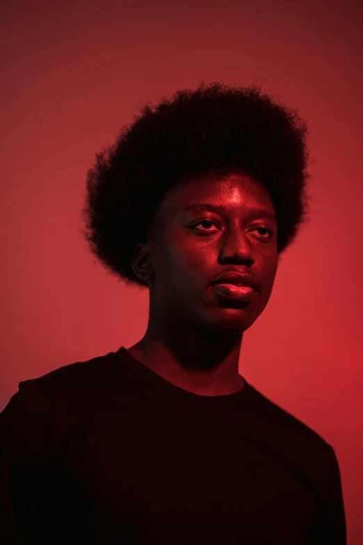 a man standing in front of a red wall, an album cover, by Lily Delissa Joseph, standing with a black background, androgynous face, ( ( dark skin ) ), red monochrome