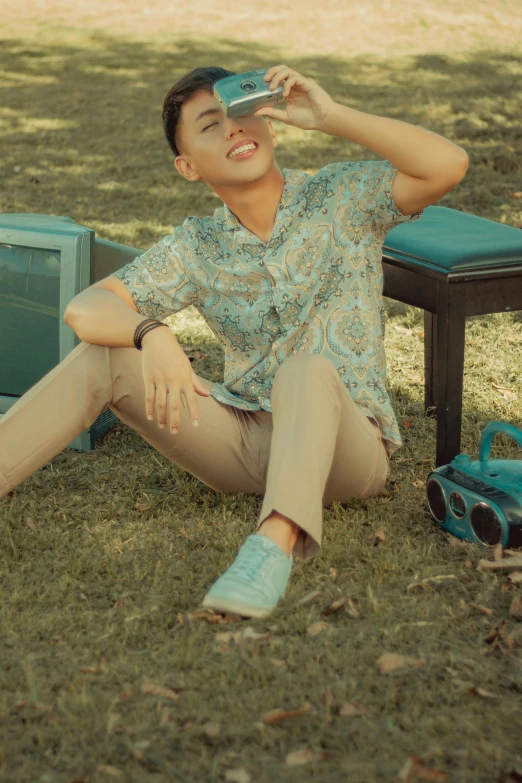 a man sitting on the ground next to a television, inspired by Rudy Siswanto, trending on pexels, patterned clothing, brown and cyan color scheme, wear's beige shirt, outdoor photo