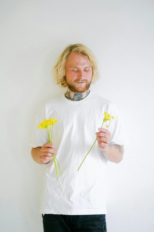 a man standing in front of a white wall holding flowers, an album cover, inspired by Lasar Segall, unsplash, dressed in a white t-shirt, medium yellow blond hair, florida man, felix englund