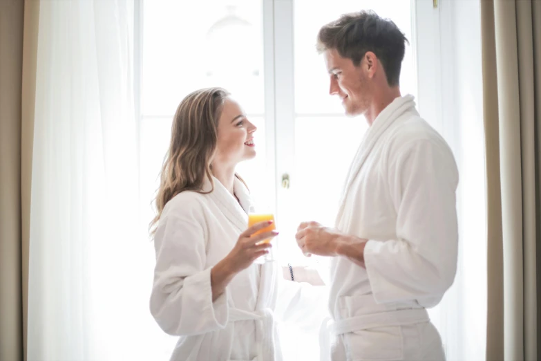 a man and a woman standing in front of a window, bathrobe, having a good time, wearing white pajamas, drinks