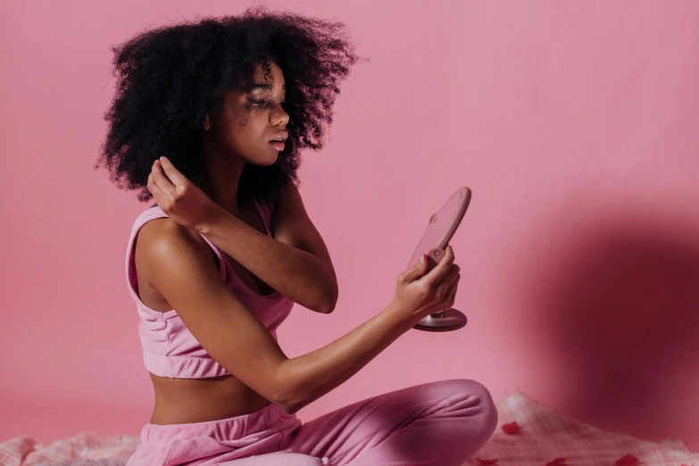 a woman sitting on a bed holding a cell phone, trending on pexels, happening, tight gigantic pink curls, looking into a mirror, focused on her neck, dark skin tone