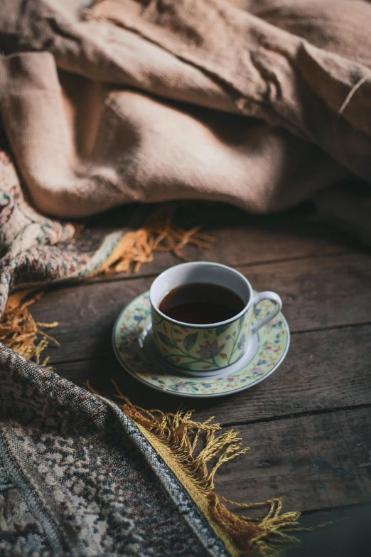 a cup of coffee sitting on top of a wooden table, a cross stitch, inspired by Elsa Bleda, trending on unsplash, renaissance, covered with blanket, secret tea society, brown clothes, promo image