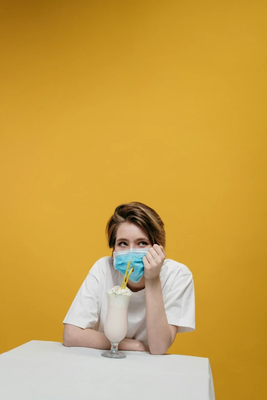 a woman sitting at a table with a mask on her face, trending on pexels, yellow backdrop, sick, ready to eat, college