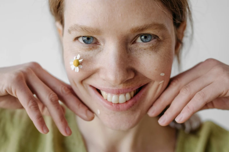 a close up of a woman with a flower on her face, trending on pexels, chamomile, smiling down from above, skincare, manuka
