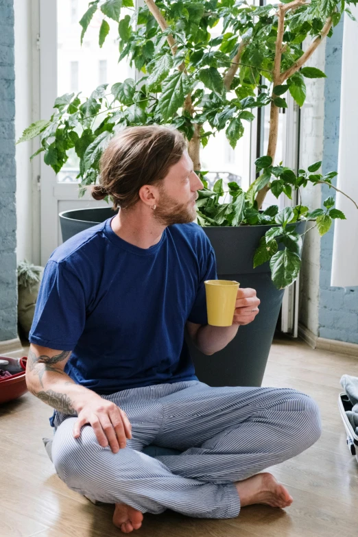a man sitting on the floor with a cup of coffee, filled with plants, profile image, wearing pants and a t-shirt, felix englund
