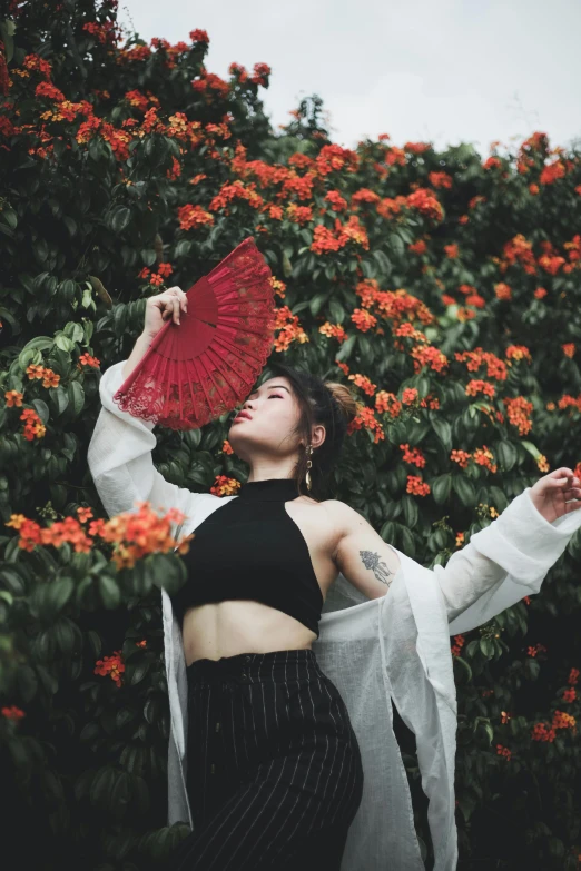 a woman standing in front of a bush holding a red fan, an album cover, inspired by Gao Xiang, unsplash contest winner, classic dancer striking a pose, flowers in her dark hair, wearing crop top, androgynous person