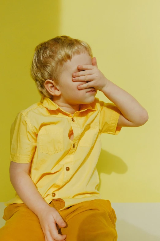a little boy sitting on the floor covering his eyes, by Lee Gatch, yellow backdrop, hand on cheek, spying discretly, partly sunny