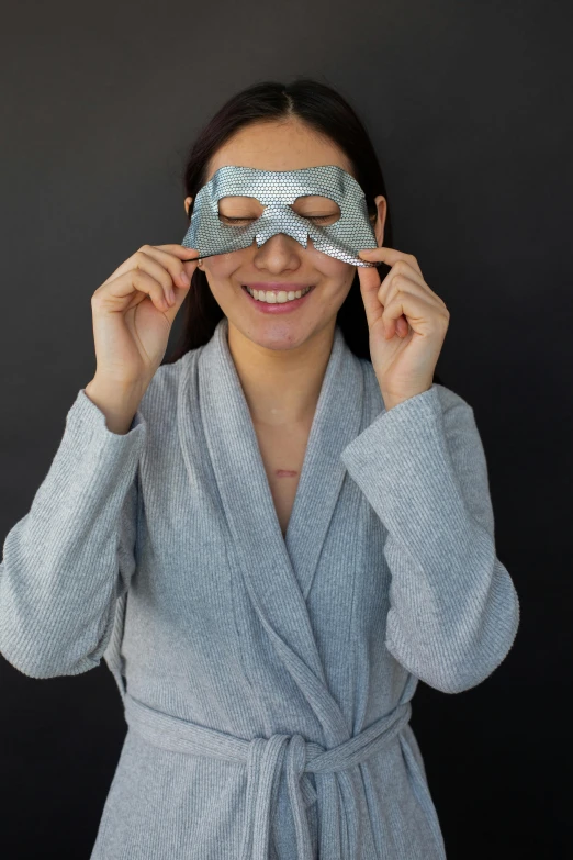 a woman in a robe holding a pair of blindfolds, featured on reddit, skin reflective metallic, on a gray background, skimask, moonlight grey