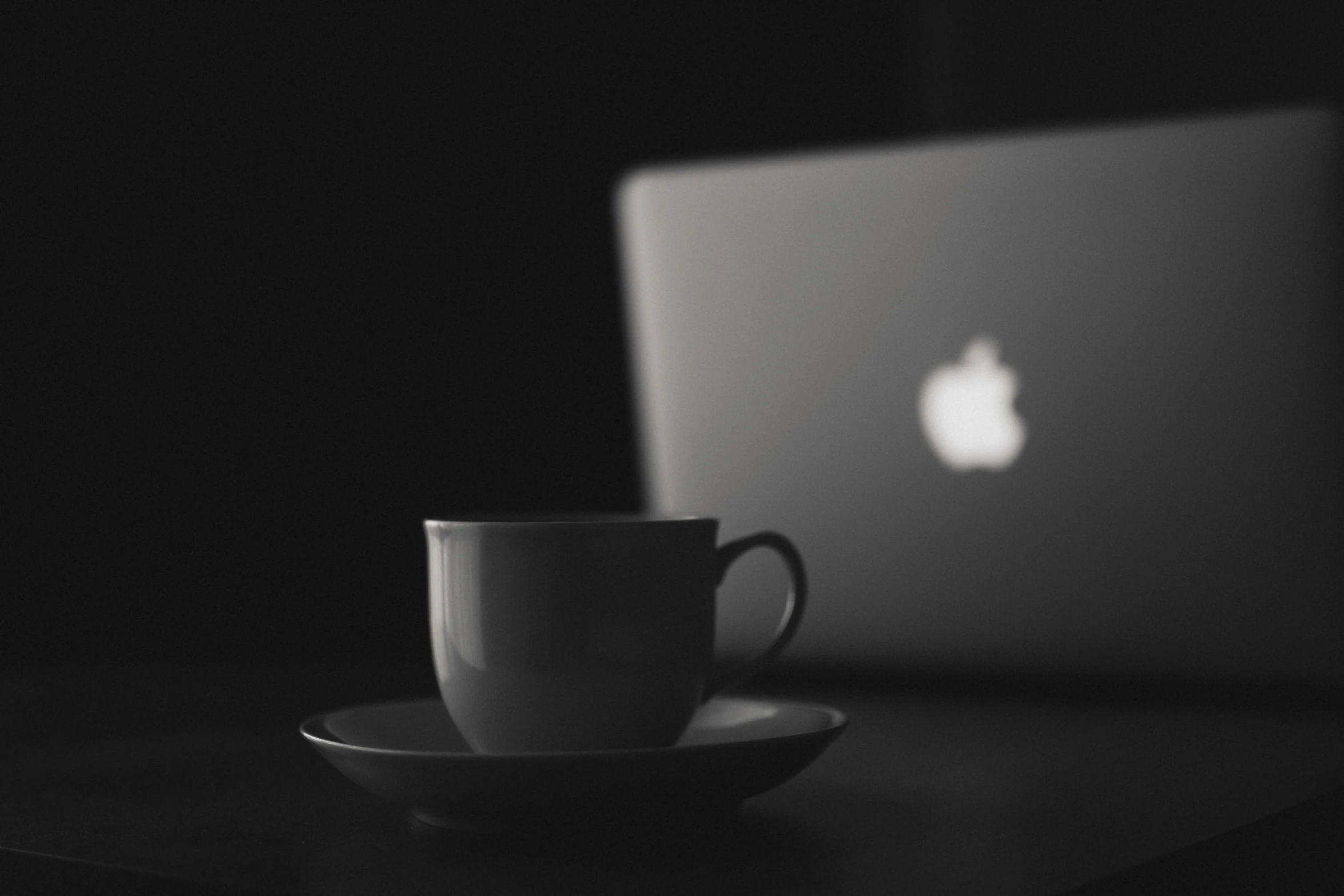 a cup sitting on top of a saucer next to a laptop, a black and white photo, by Romain brook, unsplash, apple logo, 4 k hd wallpapear, black silhouette, blurry image