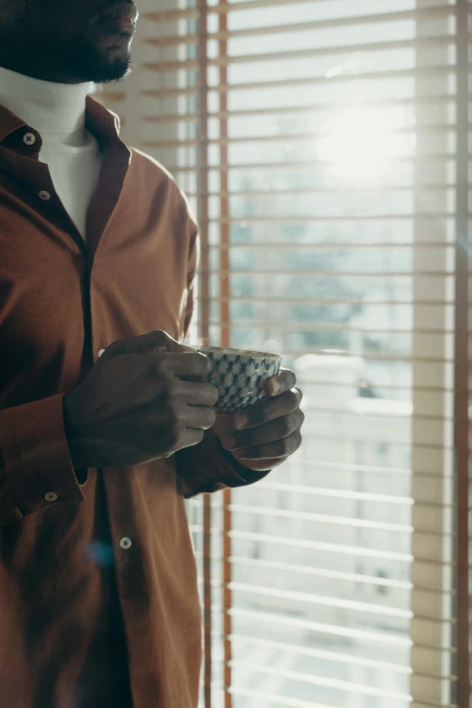a man standing in front of a window holding a cup, trending on unsplash, brown clothes, softly backlit, partially cupping her hands, blind brown man